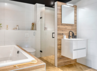 Moderne badkamer met houten accenten, witte tegels, douchecabine met glazen deur, vrijstaand bad en wit wastafelmeubel met zwarte kraan.